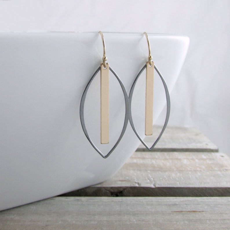 May include: A pair of gold and silver-toned earrings. Each earring features a gold vertical bar suspended within a silver-colored, leaf-shaped frame. The earrings hang from gold-colored ear wires. The background is a white surface and a wooden surface.