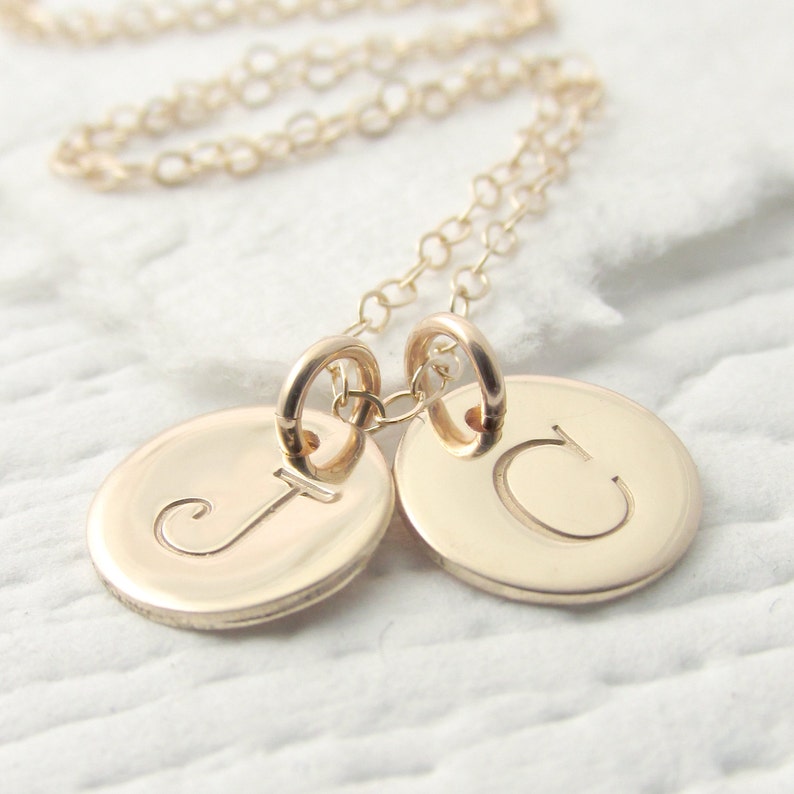 Puede incluir: Collar de oro con dos colgantes redondos, cada uno con una letra grabada. Las letras son J y C.