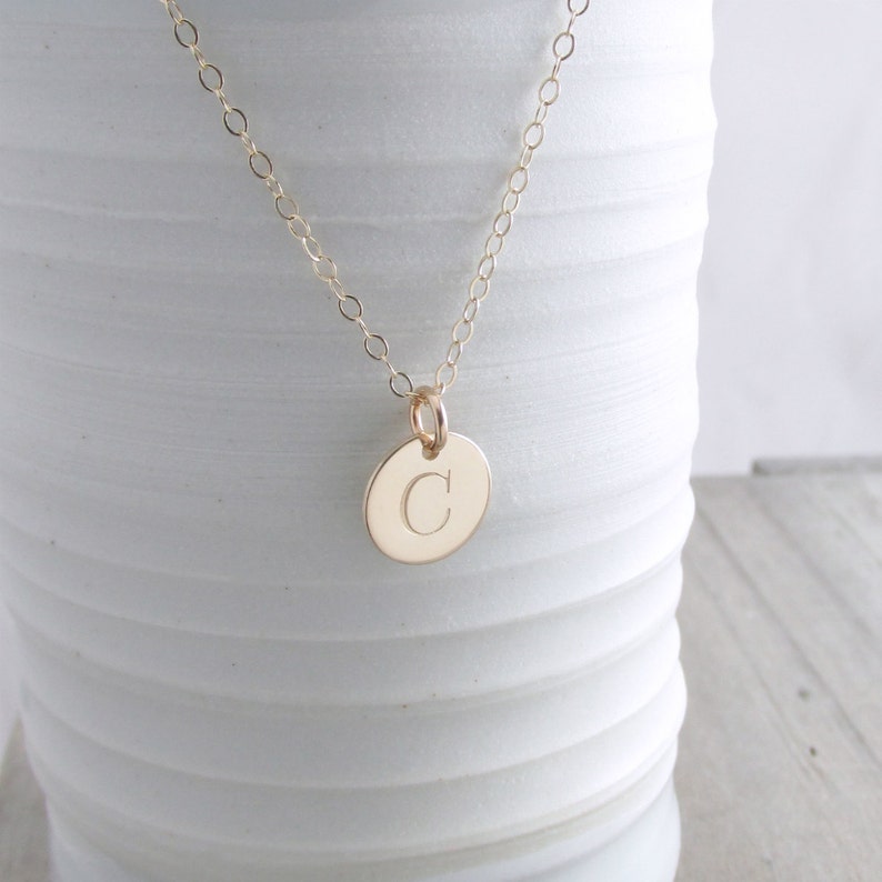 Puede incluir: Un collar de cadena de oro con un peque&ntilde;o colgante redondo con la letra "C" grabada.