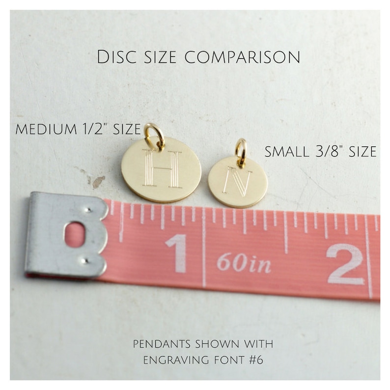 May include: Two gold-colored metal discs with engraved letters, "H" and "N", are shown next to a pink measuring tape. The tape reads "1 60in 2". The text "MEDIUM 1/2" SIZE" is above the "H" disc and "SMALL 3/8" SIZE" is above the "N" disc. The text "PENDANTS SHOWN WITH ENGRAVING FONT #6" is below the tape.