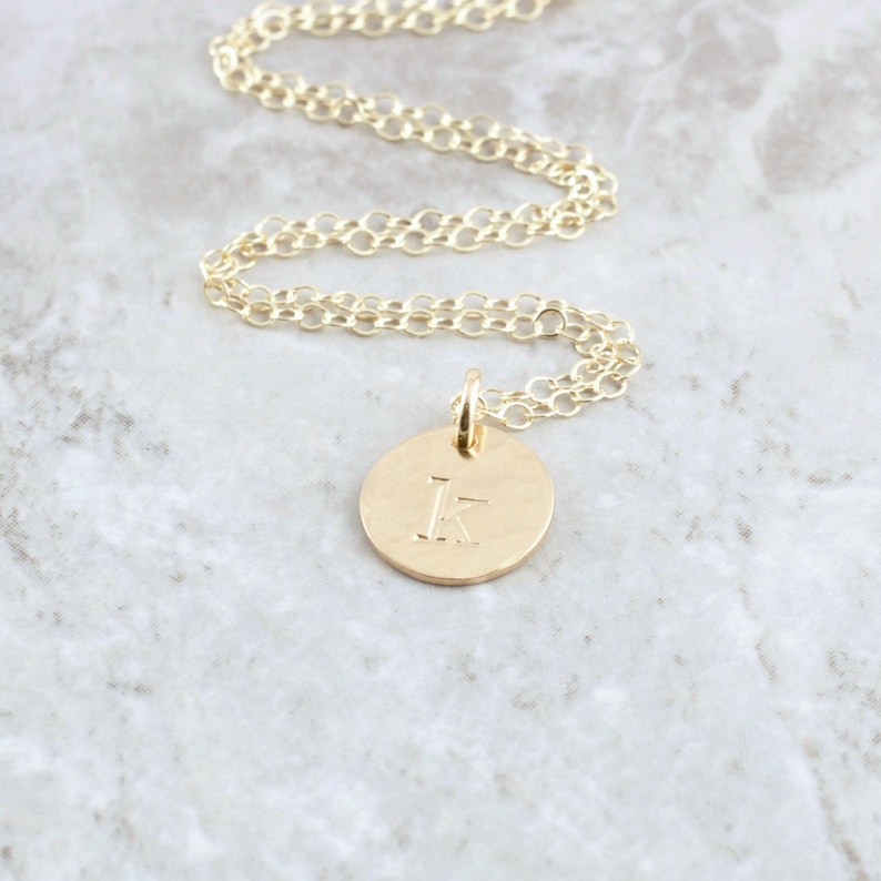 May include: A gold chain necklace with a small, round pendant featuring the letter "K" engraved on it.