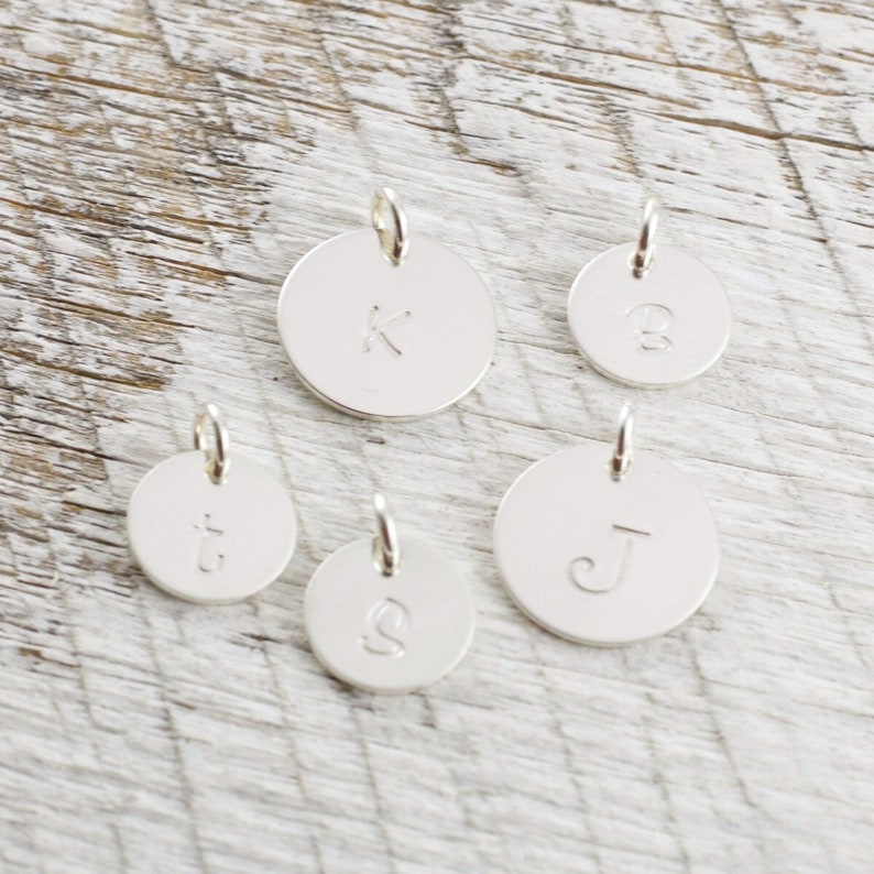 May include: Five silver charms with different letters on them. The letters are K, B, t, J, and S. The charms are on a light wood background.
