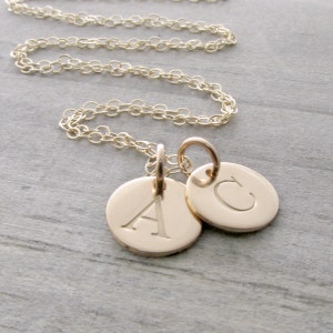 Puede incluir: Collar de cadena de oro con dos colgantes redondos, uno con la letra 'A' y el otro con la letra 'C'.