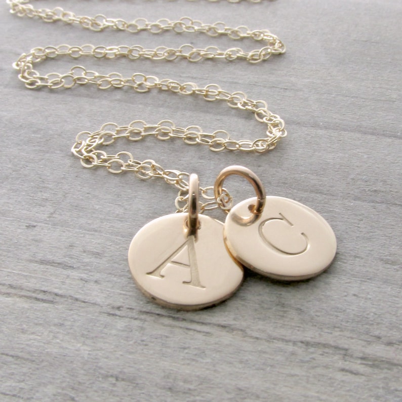 Puede incluir: Collar de cadena de oro con dos colgantes redondos, uno con la letra 'A' y el otro con la letra 'C'.