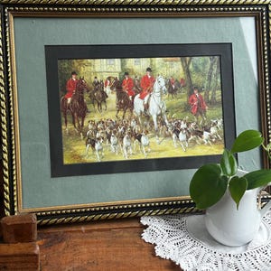 May include: A framed print depicting a fox hunt scene with riders in red coats on horseback accompanied by a pack of hounds. The artwork is enclosed in a black and gold rope-textured frame with a sage green mat and a black inner border.