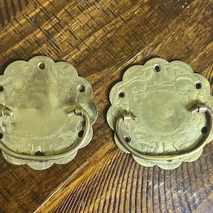 May include: Two circular brass drawer pulls featuring a scalloped edge and a central drop handle. The metal plates are engraved with intricate floral and geometric patterns, including a Greek key border design and four small mounting holes.