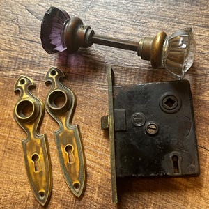Puede incluir: Un juego de herrajes antiguos para puertas que incluye un par de pomos de cristal facetado, uno morado y otro transparente, dos placas de latón con bocallaves y una caja de cerradura de embutir de metal negro con un orificio cuadrado.