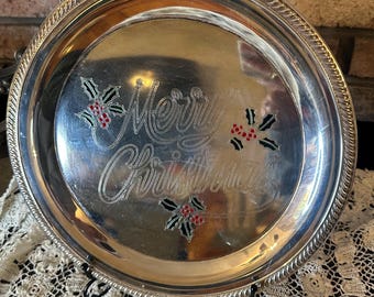 May include: A round silver-toned metal tray featuring an engraved script that reads Merry Christmas. The design includes three clusters of holly leaves with red berries. The tray has a decorative rope-style border along the outer edge.