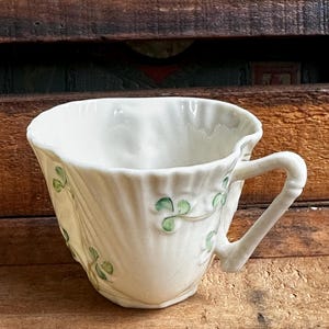 May include: A cream-colored ceramic teacup featuring a scalloped, shell-like rim and a textured, ribbed body. The exterior is decorated with delicate, hand-painted green shamrock motifs and thin vines. It includes a sturdy, angular handle.