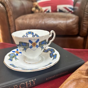 May include: A white porcelain teacup and saucer set featuring a gold rim, a blue and green tartan ribbon pattern, and a central crest with a red lion emblem. The teacup has a curved handle and sits on a matching saucer with the same decorative motif.
