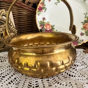 May include: A round brass basket with a polished finish and a textured exterior featuring a woven-like pattern. The container has a sturdy, arched handle attached to the sides with metal rivets, creating a functional and decorative vessel.