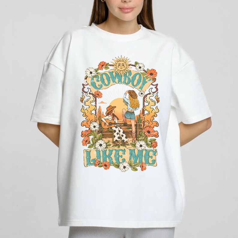 May include: A white short-sleeve t-shirt featuring a graphic print of two figures in a desert landscape with the text COWBOY LIKE ME. The design includes a sun, cacti, flowers, and flames in orange, yellow, and teal colors surrounding the central scene.