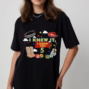 Puede incluir: Una camiseta negra con un estampado gr&aacute;fico que incluye el texto I KNEW IT, I KNEW YOU 5. El dise&ntilde;o presenta una pulsera de cuentas, un sombrero de vaquero rojo, una guitarra, botas de vaquero y bloques de letras coloridos con las letras T, TS y S.