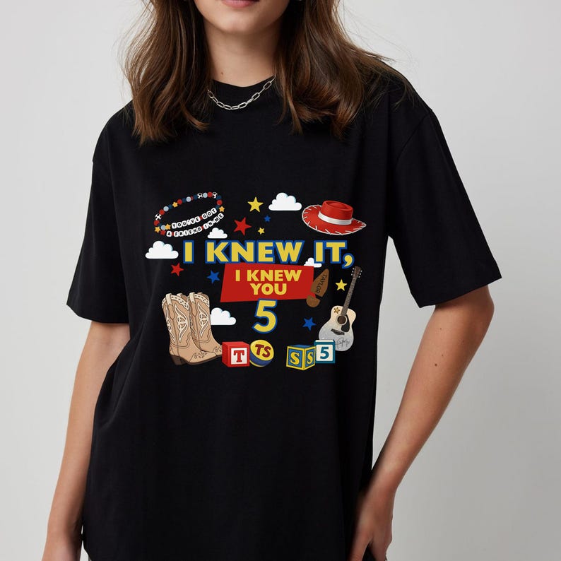 Puede incluir: Una camiseta negra con un estampado gr&aacute;fico que incluye el texto I KNEW IT, I KNEW YOU 5. El dise&ntilde;o presenta una pulsera de cuentas, un sombrero de vaquero rojo, una guitarra, botas de vaquero y bloques de letras coloridos con las letras T, TS y S.