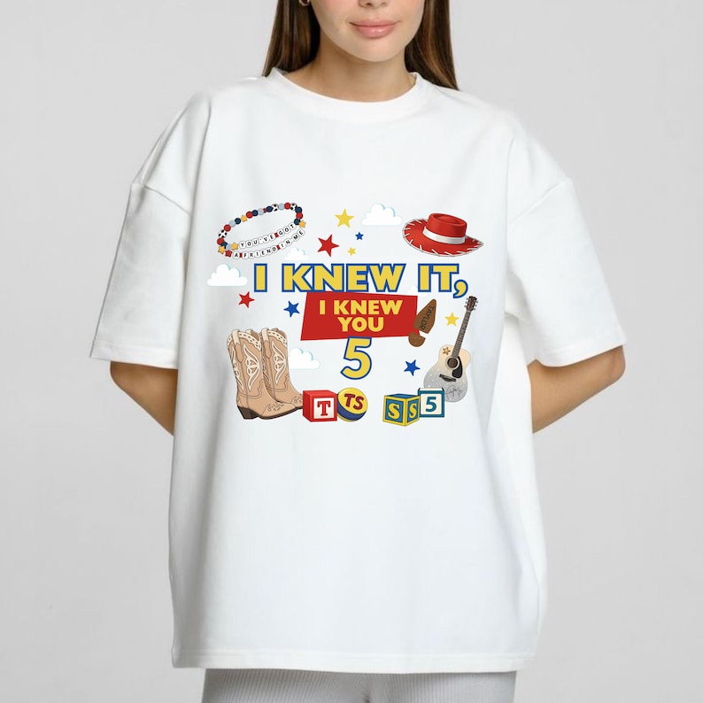 Puede incluir: Una camiseta blanca de manga corta con un estampado gr&aacute;fico que incluye el texto I KNEW IT, I KNEW YOU 5. El dise&ntilde;o presenta ilustraciones de una pulsera de cuentas, un sombrero de vaquero rojo, botas marrones, una guitarra y bloques.
