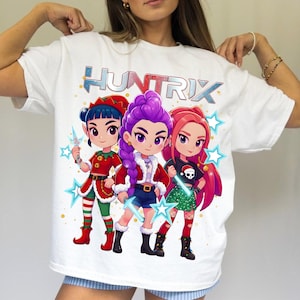 May include: White t-shirt featuring the word "HUNTRIX" in a blue and silver font above a graphic of three cartoon characters. The characters have different hair colors and outfits, with star accents.