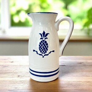 May include: White ceramic pitcher with a blue pineapple design and blue stripes. The pitcher has a handle and a spout. The background is blurred, but appears to be a window with greenery and a wooden table.