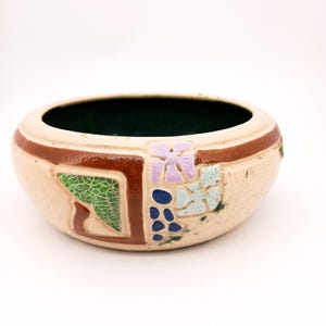 May include: A beige ceramic bowl with a dark green interior. The exterior features brown, green, blue, and lavender decorative elements. The bowl has a rounded shape and a wide opening, suitable for decorative or functional use.
