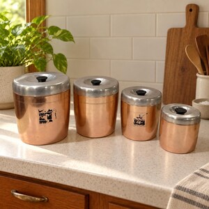 May include: A set of four cylindrical copper canisters with brushed silver lids and small black knobs. Each canister features a unique black graphic print on the front. The containers vary in size, arranged from largest to smallest in a row.