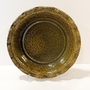 May include: A round ceramic bowl with a mottled olive green and brown glaze. The surface features a textured, speckled finish with a slightly uneven, wavy rim. The interior of the bowl is smooth and glossy, contrasting with the rustic appearance.