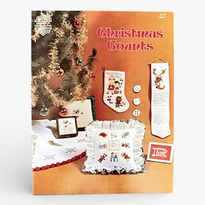 May include: A vintage Christmas-themed book cover with the title "Christmas Counts." The cover features a decorated Christmas tree, a stocking, a pillow, and other cross-stitch projects. The color palette includes red, green, and white.