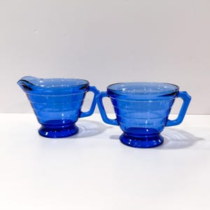 May include: A cobalt blue glass creamer and sugar bowl set. The creamer features a pouring spout and a single handle, while the sugar bowl has two handles. Both pieces are made of translucent blue glass with horizontal ribbed detailing.