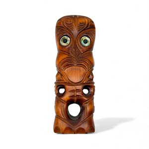 May include: A carved wooden tiki statue with a brown finish. The tiki features detailed carvings, including facial features and patterns. The eyes are inlaid with a light green and white material. The statue has a rectangular shape and stands upright.