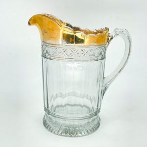 May include: A clear glass pitcher with a gold-colored rim and handle. The pitcher has a ribbed design on the body and a decorative pattern near the top. The handle is clear and curved.