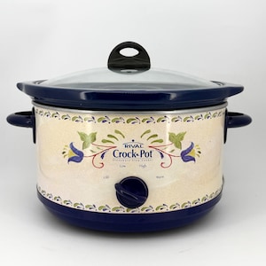 May include: A Rival Crock-Pot stoneware slow cooker with a dark blue base, lid, and handles. The cream-colored exterior features a floral pattern with blue and green accents. The front displays the text Rival Crock-Pot, Low, High, Off, and Warm.