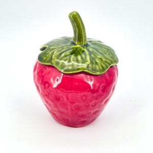 May include: A ceramic strawberry-shaped container with a green leafy lid and stem. The container is a vibrant red color, with a textured surface. The lid and stem are a glossy green. This decorative piece is ideal for kitchen decor.