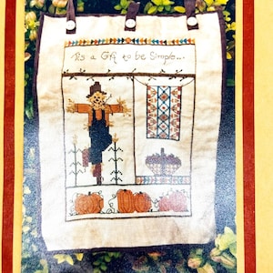 May include: A beige fabric wall hanging with a stitched design featuring a scarecrow, pumpkins, and a quilt. The text "Tis a Gift to be Simple..." is stitched across the top. Brown straps and white buttons are at the top.