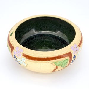 May include: A round ceramic bowl with a cream-colored exterior and a dark green interior. The rim features a brown band with floral designs in purple, blue, and green. The bowl has a slightly aged appearance.