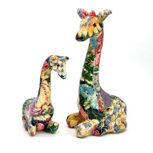 May include: Two giraffe figurines in different sizes, both featuring a vibrant, multi-colored floral pattern covering their entire bodies. The surface displays a dense arrangement of flowers in shades of yellow, blue, red, and green on a dark base.