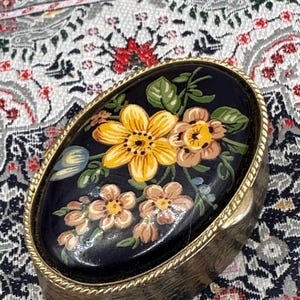 May include: Oval-shaped trinket box with a dark blue lid featuring a floral design. The flowers are yellow and peach with green leaves. The box has a gold-colored metal frame with a rope-like border.