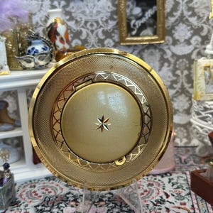 May include: A gold-toned compact mirror with a starburst design in the center. The compact has a textured border and is displayed on a clear stand. The background includes miniature framed pictures and decorative items.