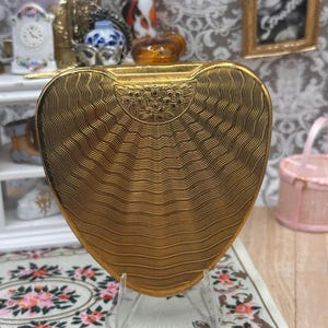 May include: A heart-shaped gold compact case featuring a textured wavy line pattern across the surface and a semi-circular decorative panel at the top engraved with a floral motif. The metal object is displayed upright on a clear plastic stand.