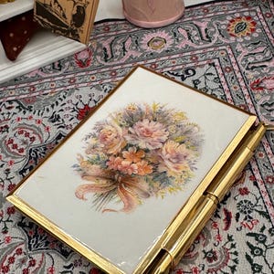 May include: A gold-toned compact with a floral bouquet design on a white background. The bouquet features pink, peach, and yellow flowers with a ribbon. The compact is rectangular with rounded corners and a pen attached.