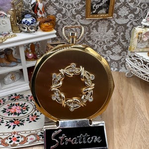 Pode incluir: Um espelho compacto redondo em tom dourado da marca Stratton com um adorno central em forma de coroa cravejado com strass transparentes. O compacto tem um acabamento metálico e uma argola superior, sobre um suporte preto com o nome Stratton.