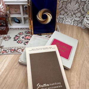 May include: A rectangular blue and gold compact with a central gold spiral design, accompanied by a gold pen. Below are two paper pads, one with a pink label reading Basildon Bond and another with a brown label reading Stratton matching handbag accessories.