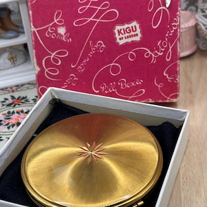 May include: A round gold-tone metal compact mirror with a starburst engraving in the centre, resting in a white box with a black velvet lining. Behind it stands a pink cardboard box featuring white cursive text and the KIGU OF LONDON logo.