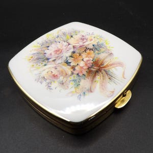 Op de afbeelding: Een vierkante, witte compactpoederdoos met een goudkleurige rand. De deksel heeft een bloemenboeketontwerp in pastelkleuren, waaronder roze, perzik en gele bloemen, met een bruin lint. De compactpoederdoos is gesloten met een gouden sluiting.