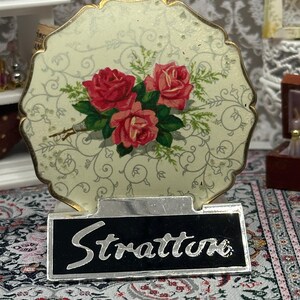 May include: Vintage Stratton compact mirror with a floral design featuring red roses and green leaves on a cream background. The compact has a gold-colored trim and the name "Stratton" is displayed on a black and silver base.
