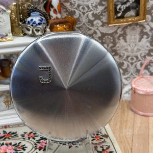 May include: A round, silver-toned metal compact case featuring a radial, sunburst-style engraved texture across its surface. The front of the compact is adorned with a single, dark-colored, capital letter J, which is embellished with small, dark stones.