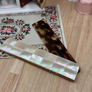 May include: A rectangular hair comb with a tortoiseshell pattern and a matching case featuring a grid of iridescent white mother-of-pearl squares. The case has a thin gold-toned metal border framing the pearlescent inlay on the front surface.