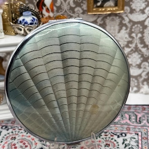 May include: A round, silver-toned compact with a fan-shaped, textured design in shades of green and silver. The compact is displayed on a clear stand, showcasing its intricate details and vintage aesthetic. A classic accessory.