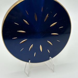 Op de afbeelding: Een vintage, ronde compacte poederdoos met een marineblauwe emaille bovenkant en goudkleurige metalen rand. De bovenkant heeft een zonnestraalontwerp van goudkleurige, bladvormige accenten. De compacte poederdoos wordt weergegeven op een heldere acryl standaard.