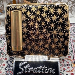 May include: A vintage Stratton compact mirror. The black compact features a gold snowflake pattern and a gold lipstick holder. The base is silver-toned with the cursive name "Stratton".