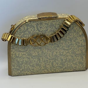 Könnte beinhalten: Vintage cremefarbene Handtasche mit floralem Muster und goldfarbener Hardware. Die Tasche hat einen dekorativen goldenen Verschluss und einen segmentierten goldenen Griff. Der Verschluss ist ein kompliziertes Knotendesign. Die Tasche hat eine rechteckige Form mit abgerundeten Kanten.
