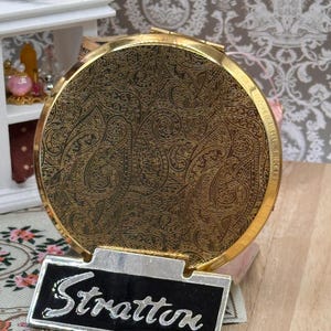 May include: A round gold-tone metal compact mirror featuring an intricate black engraved paisley pattern on the lid. The compact is displayed on a rectangular silver-tone metal stand with a black insert that has the word Stratton written in silver.