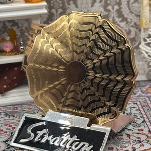 May include: A vintage Stratton compact mirror with a gold-tone, fan-shaped design and black accents. The compact features a black and silver nameplate with the name "Stratton" in cursive script. The compact is displayed on a patterned surface.